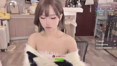 Media: A video of a young Asian woman with fair skin and shoulder-length brown hair, wearing a black and white fur-trimmed off-shoulder top, sitting in a kitchen with a virtual background.