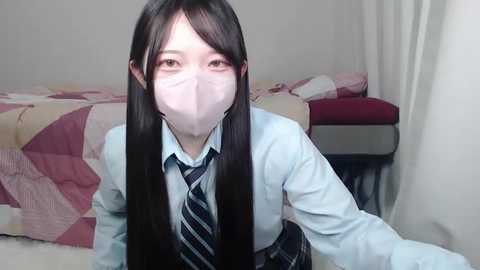 Media: Video of a young Asian woman with long black hair, wearing a light blue shirt, dark tie, and face mask, kneeling on a bed with a pink and red quilt.