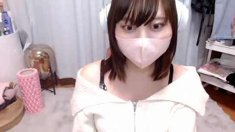 Media: Video of a young Asian woman with light skin, shoulder-length brown hair, and large headphones, wearing a white mask, white hoodie, and black bra. Background includes white curtains, a pink candle, and a white table.