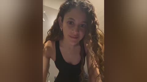 Media: Video of a young woman with curly brown hair, wearing a black tank top, standing indoors with a blurred background.