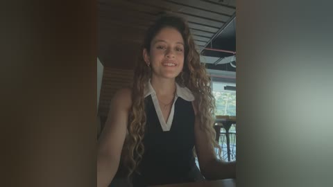 Media: A video of a smiling, young woman with long, wavy, brown hair in a dark sleeveless top, standing in a dimly lit, modern cafe with wooden ceiling beams and glass windows.