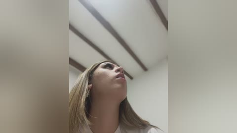 Media: Video of a young Caucasian woman with straight, blonde hair, fair skin, and a white shirt, looking upward. She stands in a modern, minimalist room with white walls and wooden ceiling beams.