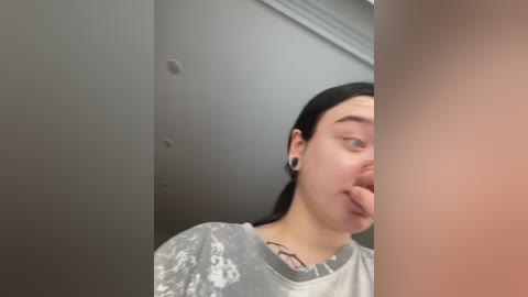 Media: Video of a young woman with fair skin, black hair, and a grey shirt, sticking her tongue out. Background features a grey wall with a hole and a beige wall.