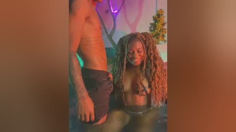 Media: A video of a shirtless, tattooed man in black shorts standing next to a woman with long, curly brown hair, partially submerged in water. The background features colorful, abstract wall art and a potted plant.