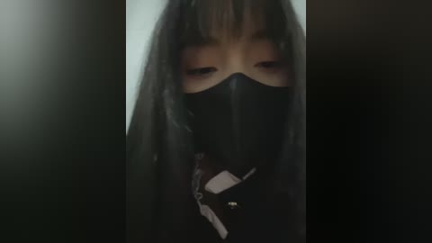 Media: Video of a person with long black hair, wearing a black mask, standing in a dimly lit room. The image is dark and mysterious, with minimal background detail.