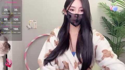 Media: Video of an Asian woman with long black hair, wearing a white robe with bear patterns, a black mask, and a blue top. Background includes a green plant and a pink hula hoop.