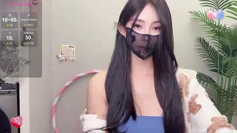 Media: Video of an Asian woman with long black hair, wearing a blue tank top and a black mask, standing in a room with a hula hoop, a plant, and a wall-mounted thermometer.