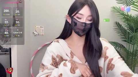 Media: Video of an East Asian woman with long black hair, wearing a face mask, a fluffy bear-patterned pajama, and a green plant in a room.