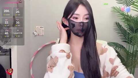 Media: Video of an East Asian woman with long black hair, wearing a face mask and a blue top with teddy bear pattern, sitting in a room with a green plant and a wall clock.