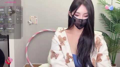 Media: Video of an East Asian woman with long black hair, wearing a mask, a light jacket with teddy bears, and a blue top, seated in a room with a thermometer, a potted plant, and a pink hoop.