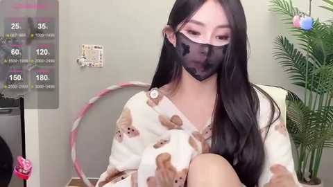 Media: Video of an Asian woman with long black hair, wearing a white pajama top with teddy bears and a black face mask, seated in a room with a potted plant and a wall-mounted thermometer.