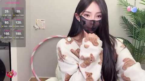 Media: Video of an East Asian woman with long black hair, wearing a white bear-patterned pajama top, black mask, and a pink hula hoop, indoors with a calendar and green plant.