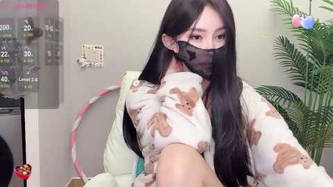 Media: Video of an Asian woman with long black hair, wearing a mask and a pajama shirt adorned with teddy bears, in an indoor setting with a potted plant and a clock on the wall.