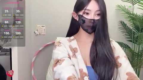 Media: Video of an East Asian woman with long black hair, wearing a beige bear-print robe, black face mask, and a blue top, in a home setting with a plant and digital display.