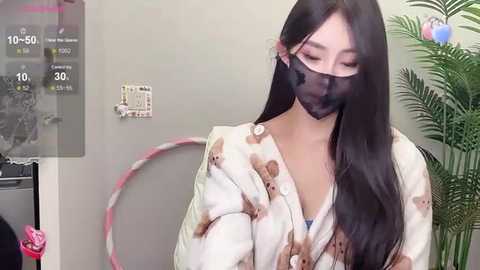 Media: Video of an Asian woman with long black hair, wearing a white blouse with brown patterns and a black mask, standing in a modern room with a green plant and a digital display showing temperature and time.