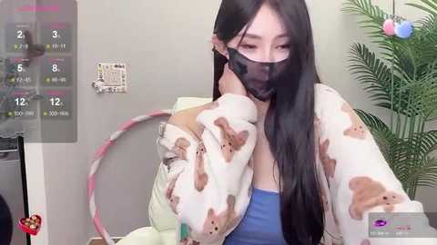 Media: A video of an East Asian woman with long black hair wearing a pink and white bunny-themed jacket, blue tank top, and black face mask, sitting in a modern room with a green plant and calendar.