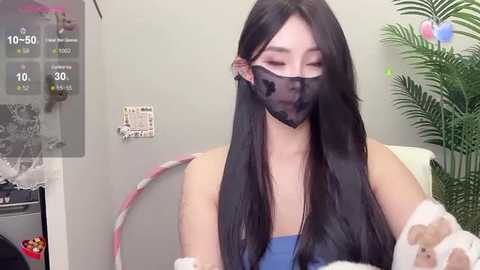 Media: Video of an East Asian woman with long black hair, wearing a black face mask and blue tank top, sitting on a white chair in a room with a green plant and gray wall.