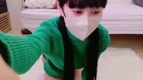 Media: Video of a young Asian woman with straight black hair and bangs, wearing a green knitted sweater, white face mask, and black gloves, taking a selfie in a brightly lit bedroom with a white bed and wooden furniture.