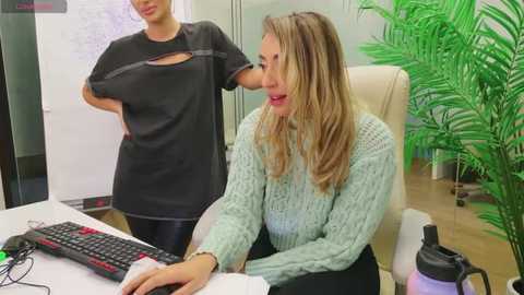 Media: Video of two women in an office setting. One wears a black shirt, the other a light blue sweater. Green plant, keyboard, and water bottle on desk.