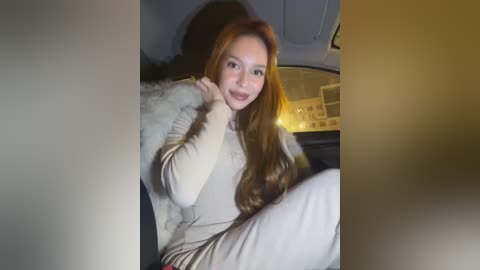 Media: Video of a fair-skinned woman with long, wavy auburn hair, wearing a beige sweater and pants, sitting in a car with a blurred background.