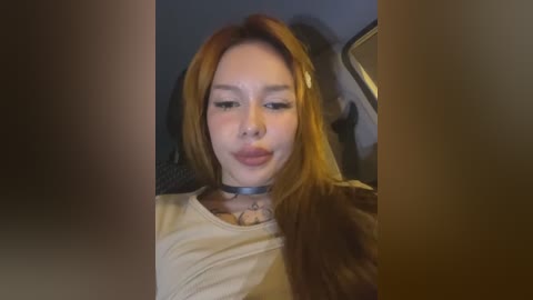 Media: A video of a young woman with long, straight auburn hair, light skin, and wearing a black choker, seated in a car, with a neutral expression.