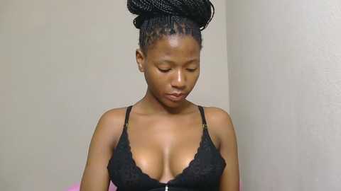 Media: Video of a young African woman with medium brown skin, wearing a black lace bralette, with intricately braided hair styled in an updo. She has a slender physique, small breasts, and is standing against a plain beige wall.