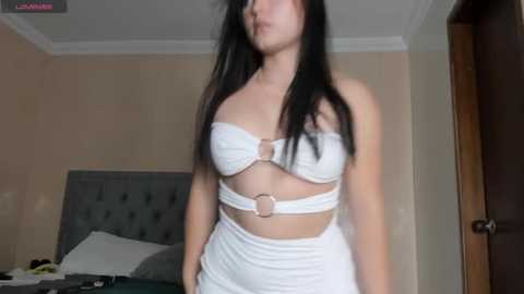 Media: Video of a young Asian woman with long black hair, wearing a white strapless top and matching skirt, standing in a simple, beige-walled bedroom with a gray upholstered bed and door in the background.