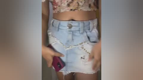 Media: Video of a woman wearing a floral crop top and distressed, light blue denim mini skirt with frayed edges, holding a red phone.