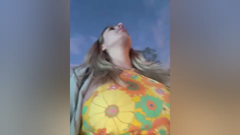 Media: Video of a woman with long blonde hair, wearing a bright yellow dress with orange and green floral patterns, looking up at a clear blue sky.