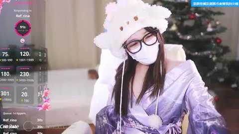 Media: Video of an East Asian woman with long black hair, wearing a white fur hat, glasses, and a white mask, sitting indoors, with a Christmas tree in the background.