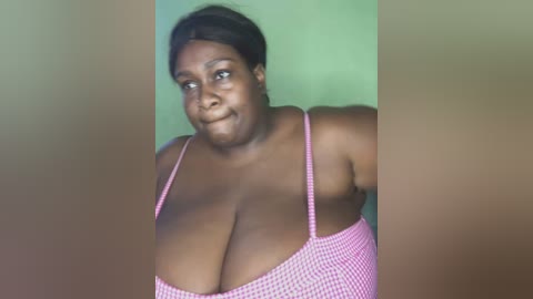 Media: A video of a plus-sized Black woman with dark skin and short hair wearing a pink and white checkered bra, smiling against a green background.