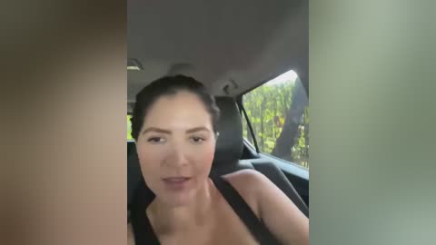 Media: Video of a woman with light skin, dark hair tied back, wearing a black top, seated in a car with greenery visible outside the window.