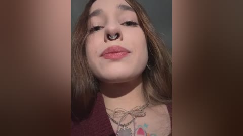 Media: Video of a young woman with pale skin, brown hair, and a septum piercing, wearing a burgundy top and a silver necklace.