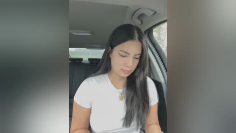 Media: Video of a young Latina woman with long black hair and fair skin, wearing a white t-shirt, seated in a car, looking down at her phone.