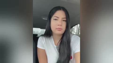 Media: Video of a young woman with long black hair, wearing a white T-shirt, sitting in a car. The background shows a blurred view of the car interior and outside.