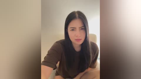 Media: Video of a young Asian woman with long black hair, wearing a brown t-shirt, sitting on a bed, looking directly at the camera with a neutral expression.