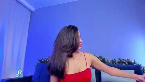 Media: A video of a young woman with long, straight black hair, wearing a red spaghetti-strap top, sitting in a dimly lit room with blue lighting, decorative plants, and a Christmas tree in the background.