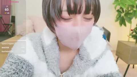 Media: Video of an Asian woman with short brown hair, wearing a white mask, lying on a bed, wearing a fluffy gray and white robe.