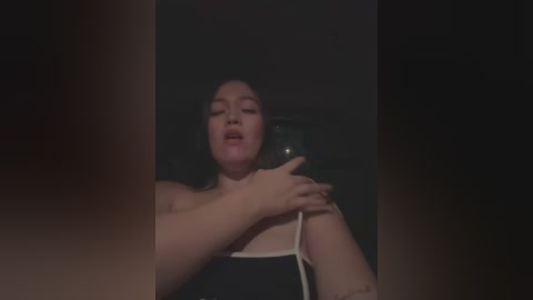 Media: Video of a plus-sized, light-skinned woman with dark hair, wearing a black top, holding a white cord, in a dimly-lit room.