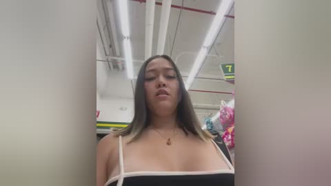 Media: Video of a young Asian woman with long, straight, black hair, wearing a low-cut black dress with white straps, standing in a brightly lit convenience store with fluorescent lights and signs.