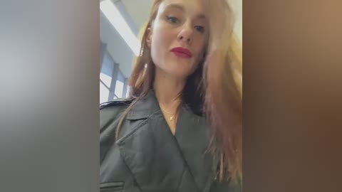 Media: Video of a fair-skinned woman with long, straight, reddish-brown hair, wearing a dark, double-breasted jacket, standing indoors with fluorescent lighting and a window in the background.