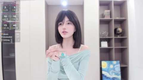 Media: Video of a young woman with straight black hair and pale skin, wearing a light blue off-shoulder top, standing in a modern room with a wall-mounted clock, a shelf with books, and a Van Gogh painting.