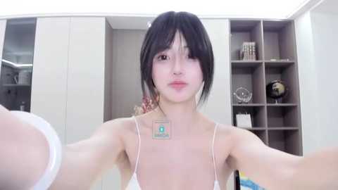 Media: Video of a young East Asian woman with short black hair and light skin, wearing a white spaghetti-strap top, taking a selfie in a modern, minimalist room with white shelves and decorative items.