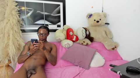 Media: Video of a nude, fit Black man with glasses, holding a phone, lying on a pink bed with plush toys, including a teddy bear and a red heart.