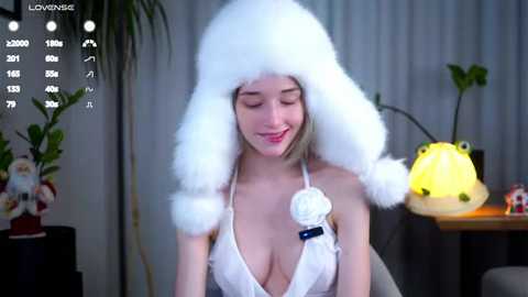 Media: Video of a smiling, fair-skinned woman with long blonde hair, wearing a fluffy white hat and a revealing white dress with a white rose. She sits in a cozy room with a lit lamp, a potted plant, and a Christmas tree in the background.