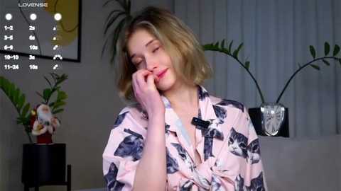 Media: Video of a blonde woman with short hair, wearing a light pink floral robe, sitting in a cozy room with potted plants and a clock.