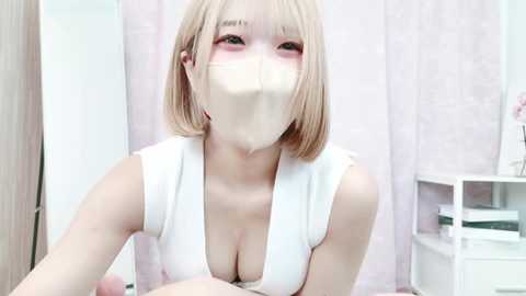 Media: Video of a young Asian woman with shoulder-length blonde hair, wearing a white sleeveless dress with a deep V-neck, revealing cleavage. She has a white face mask covering her mouth and nose, and is seated indoors against a pastel-colored backdrop.