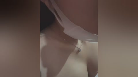 Media: A close-up video of a woman's bare chest, with a white bra strap visible, and a delicate silver necklace with a small pendant. The lighting casts soft shadows, emphasizing the smooth, pale skin.