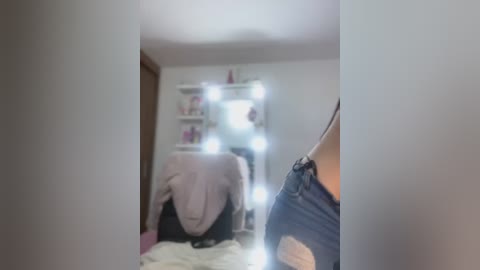 Video of a dimly lit bedroom with a white vanity mirror reflecting a woman in a black tank top and jeans, standing in front of a white shelf with pink decor.