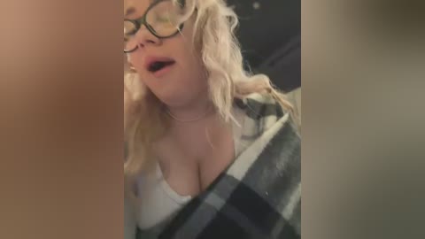 Media: Video of a blonde woman with glasses and a white shirt, wearing a black and white plaid blanket, eyes closed, mouth open in a seductive pose.